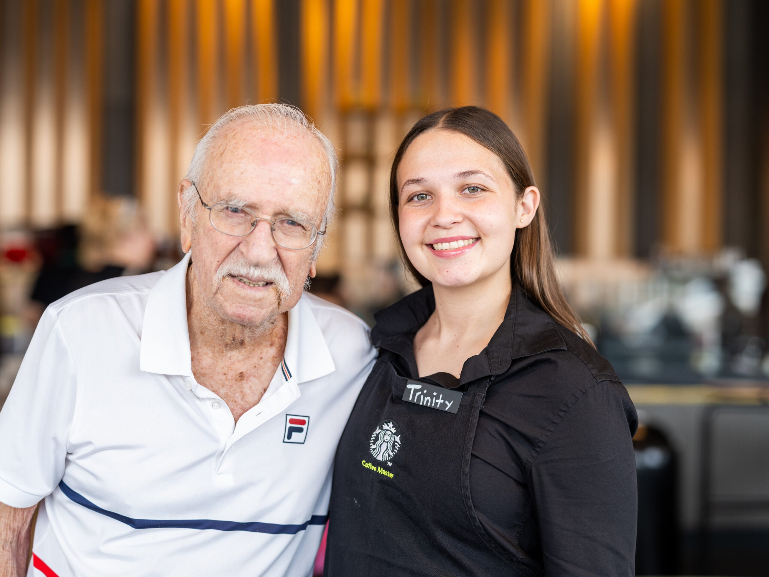 A smiling older man and a young woman pose together. The man wears glasses and a white polo shirt, while the woman, in a black apron, has a name tag. The background features warm lighting and a casual setting.