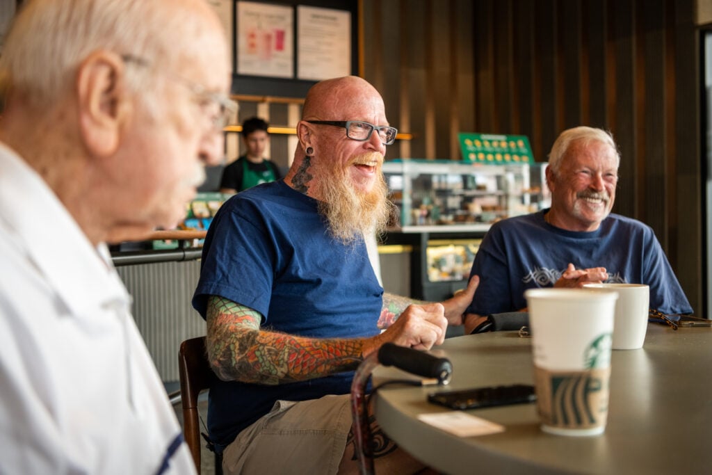 Three older men are seated at a table in a café, engaged in conversation. One man, with a bald head and a long beard, has colorful tattoos on his arms and is wearing glasses. He appears to be laughing. Another man, partially visible, has gray hair and is also smiling. A third man, in the background, is working at the counter. A Starbucks cup and a phone are on the table.