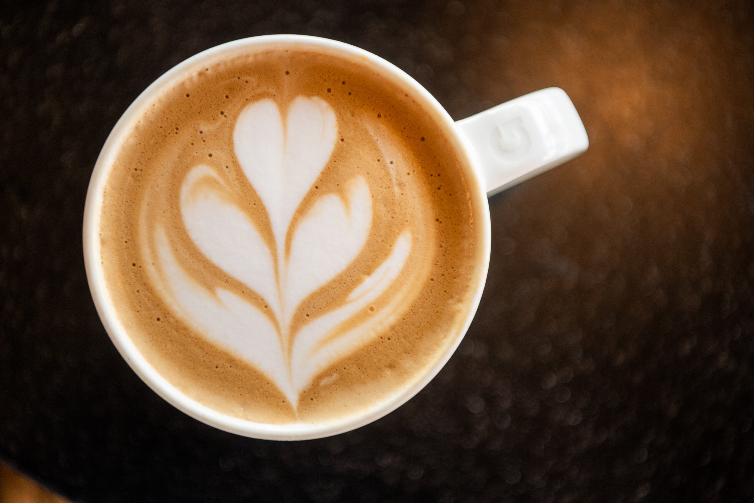A close-up of a latte in a white cup, featuring intricate heart-shaped latte art on a creamy surface, set against a dark background.