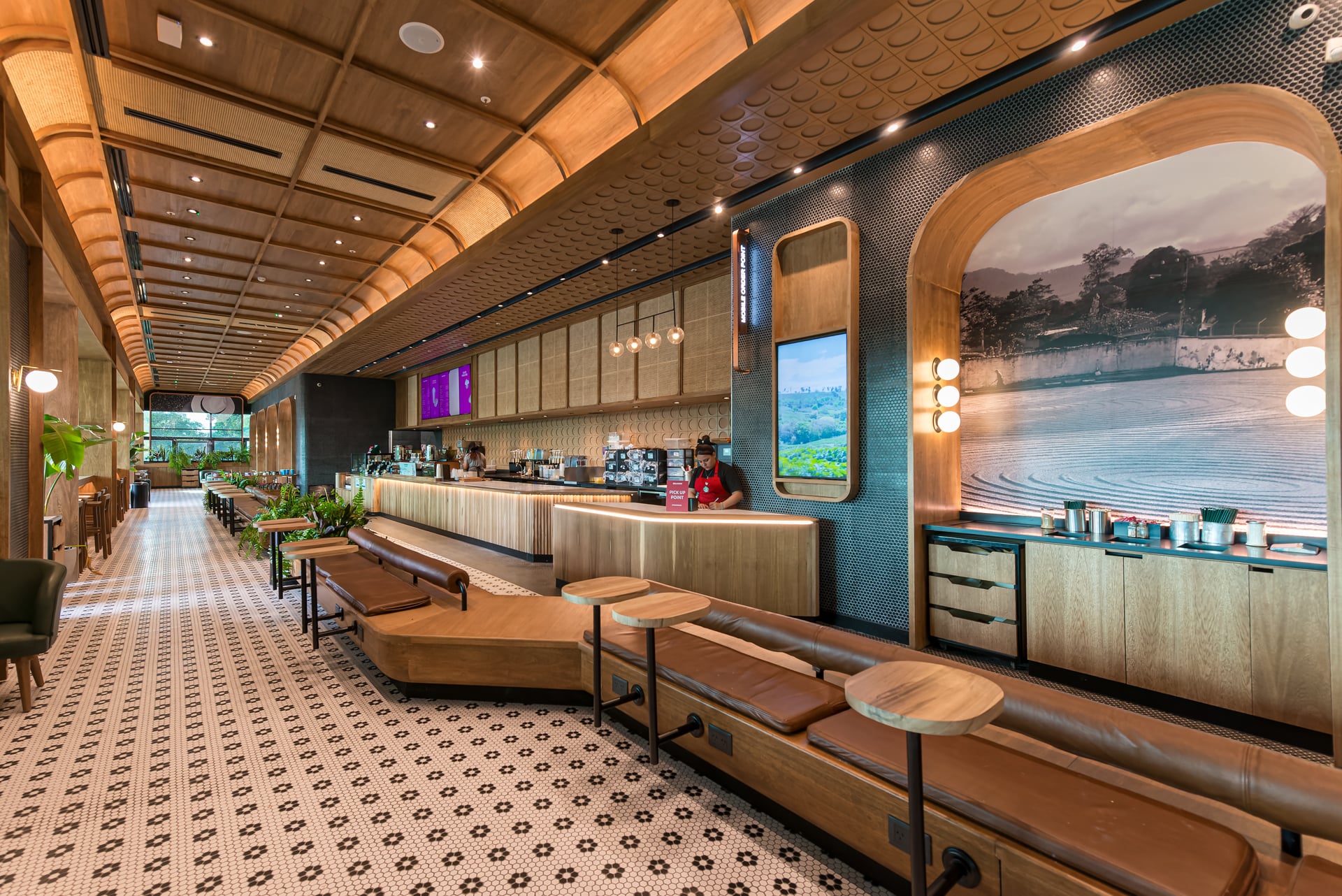 A modern café interior featuring wooden accents, a long bar with coffee equipment, and cozy seating. The floor has a patterned tile design, and large windows allow natural light.