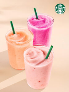 Three colorful frozen beverages are displayed in clear cups with green straws. The drinks are peach, pink, and purple, set against a light background.
