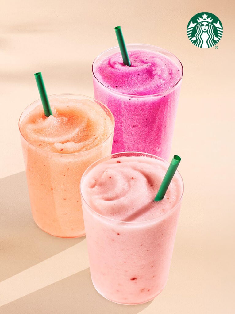 Three colorful frozen beverages are displayed in clear cups with green straws. The drinks are peach, pink, and purple, set against a light background.