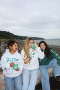 Three young women sit on a log by the water, wearing hoodies and casual jeans. One holds a drink, another a snack, and they smile.