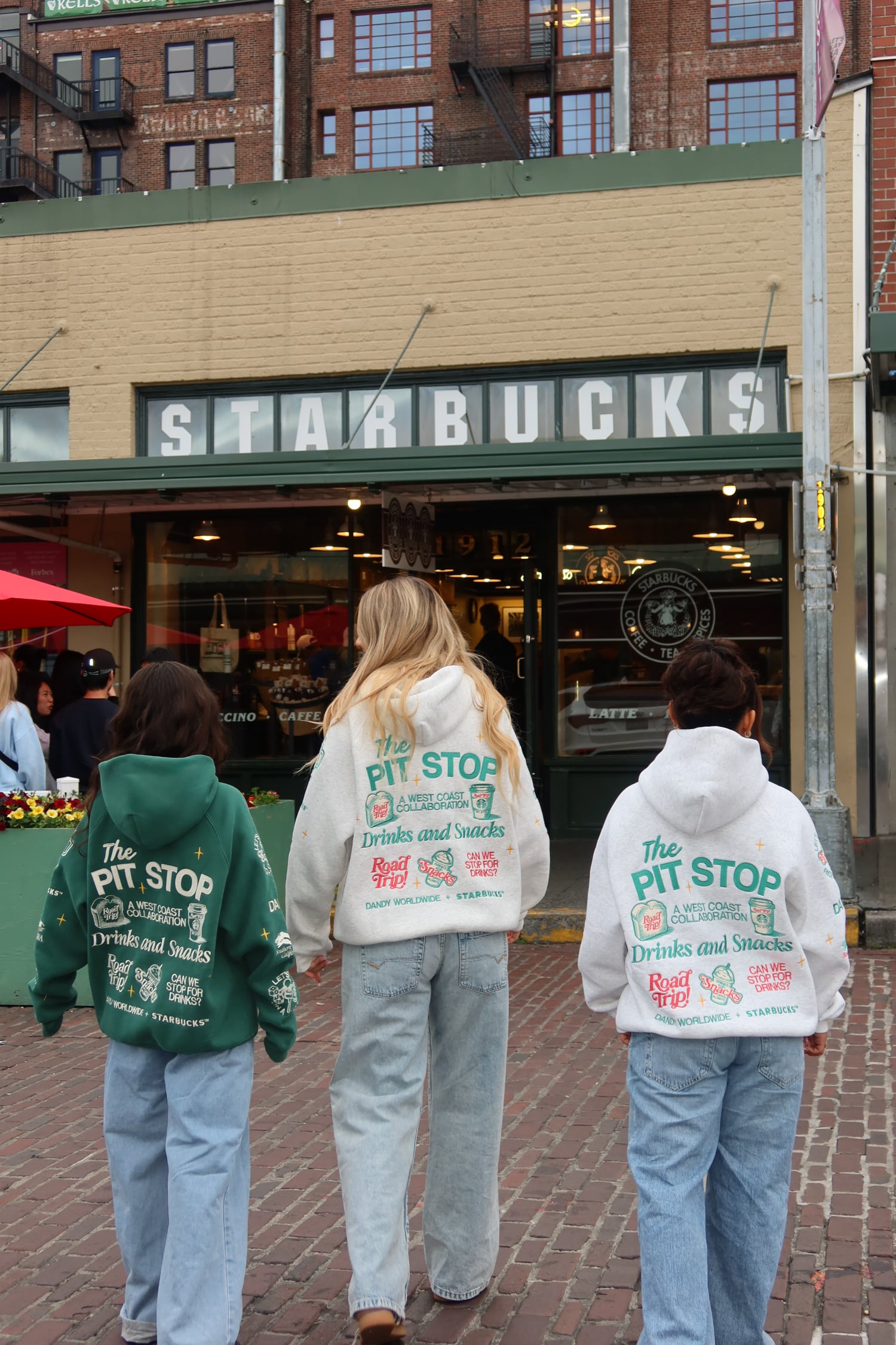 Three individuals walk towards a Starbucks, wearing colorful hoodies featuring "The Pit Stop" branding. The setting has brick pavement and a lively atmosphere.