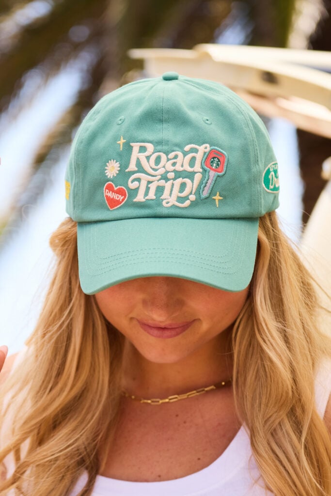 A mint green cap features the embroidered text "Road Trip!" along with colorful patches, including a heart and a key. The wearer has long, wavy hair and is dressed in a white top.