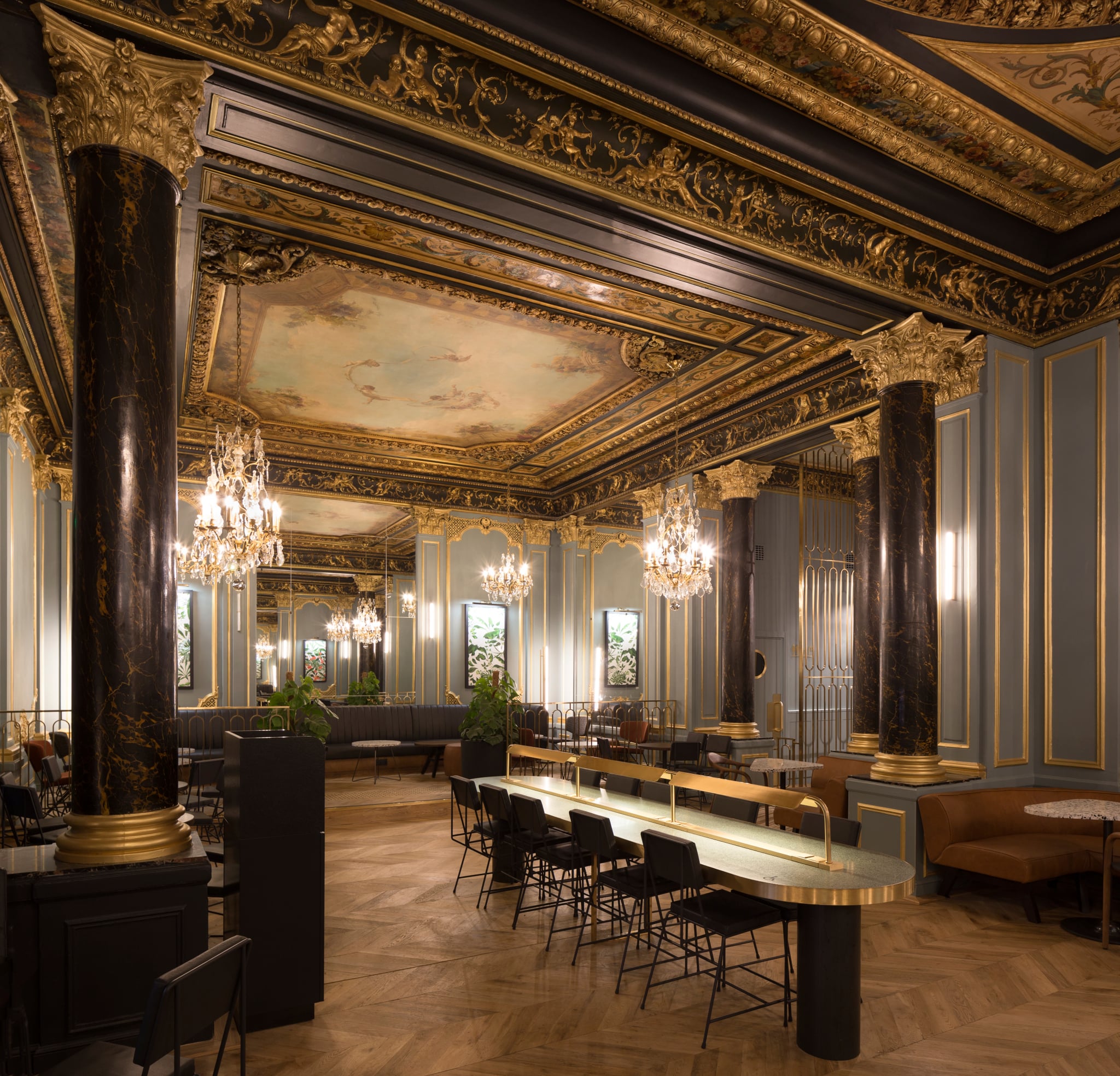 An elegant interior featuring ornate ceilings with gold detailing, black marble columns, and chandeliers. Stylish seating areas and a long bar enhance the sophisticated atmosphere.