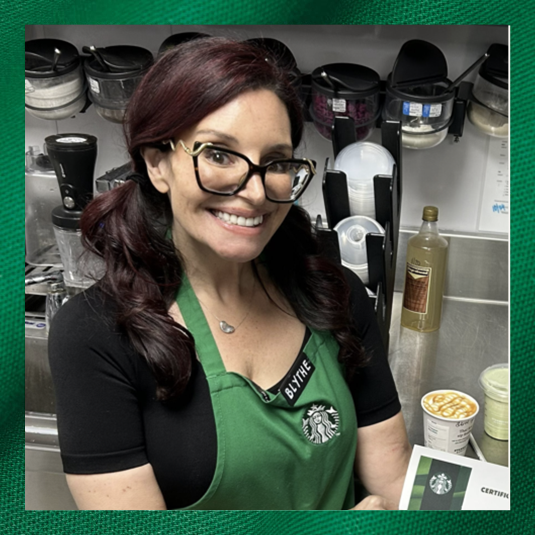 A smiling barista with long, dark hair styled in pigtails wears glasses and a green Starbucks apron. Behind her are various kitchen tools and containers. She holds a certificate and a cup with caramel drizzle.
