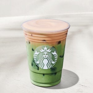 A clear cup filled with a layered drink featuring green matcha at the bottom and a peach-colored top, with ice visible throughout. The cup has a prominent logo on the front.