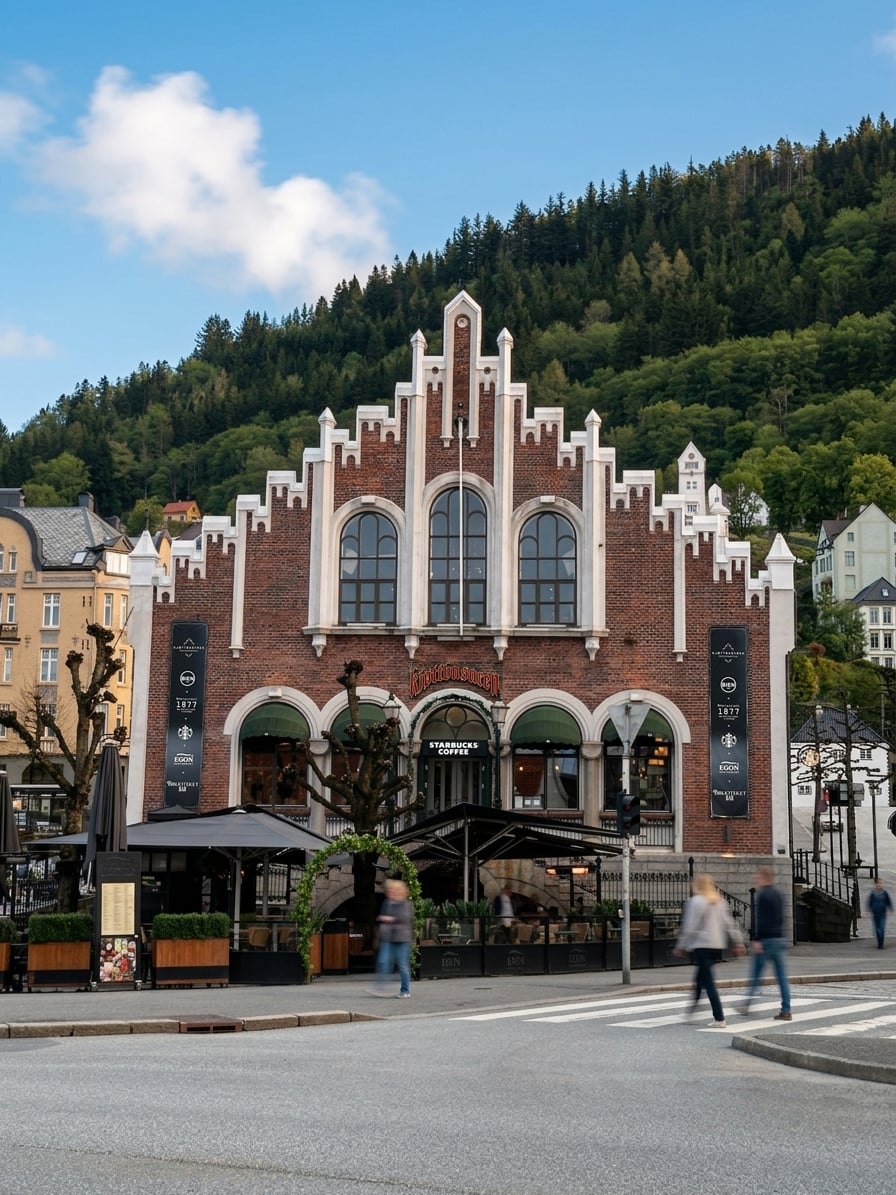A historic brick building with a distinctive gabled roof features large arched windows. Outdoor seating is visible, along with pedestrians crossing the street. Lush greenery and signage for Starbucks and a restaurant are present. The backdrop includes a forested hillside under a partly cloudy sky.