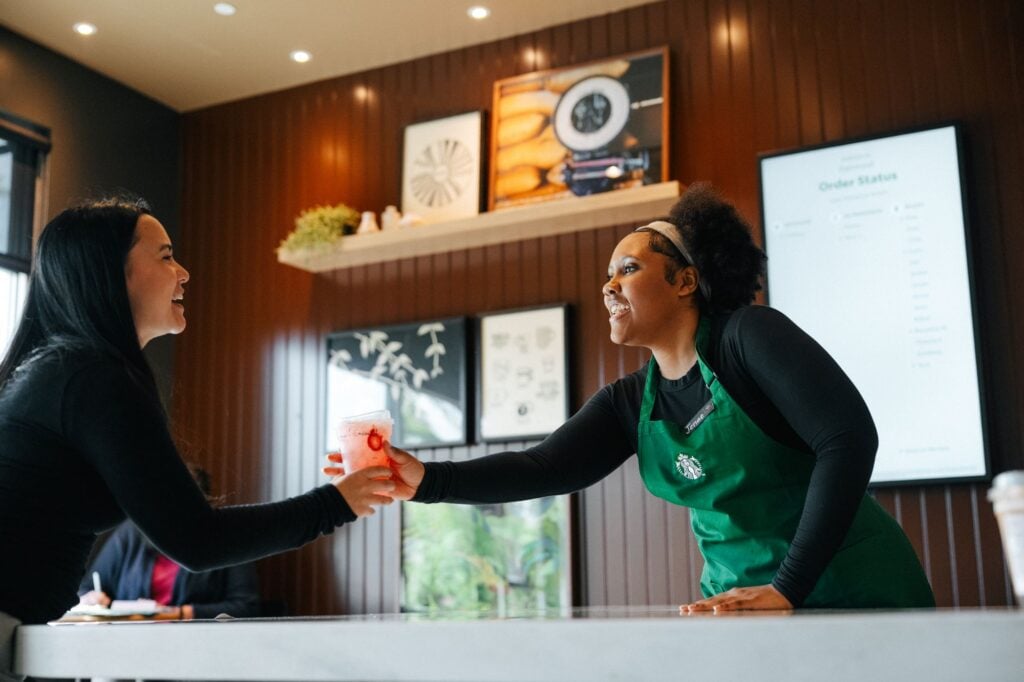 A barista in a green apron hands a drink to a smiling customer at a café. The interior features wooden walls and decorative art.