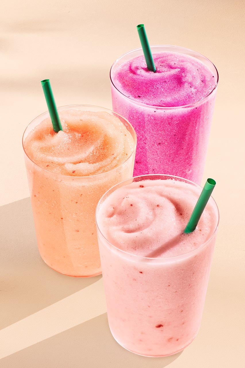 Three colorful smoothies are displayed in clear cups with green straws. The flavors appear to be peach, pink, and a vibrant purple.