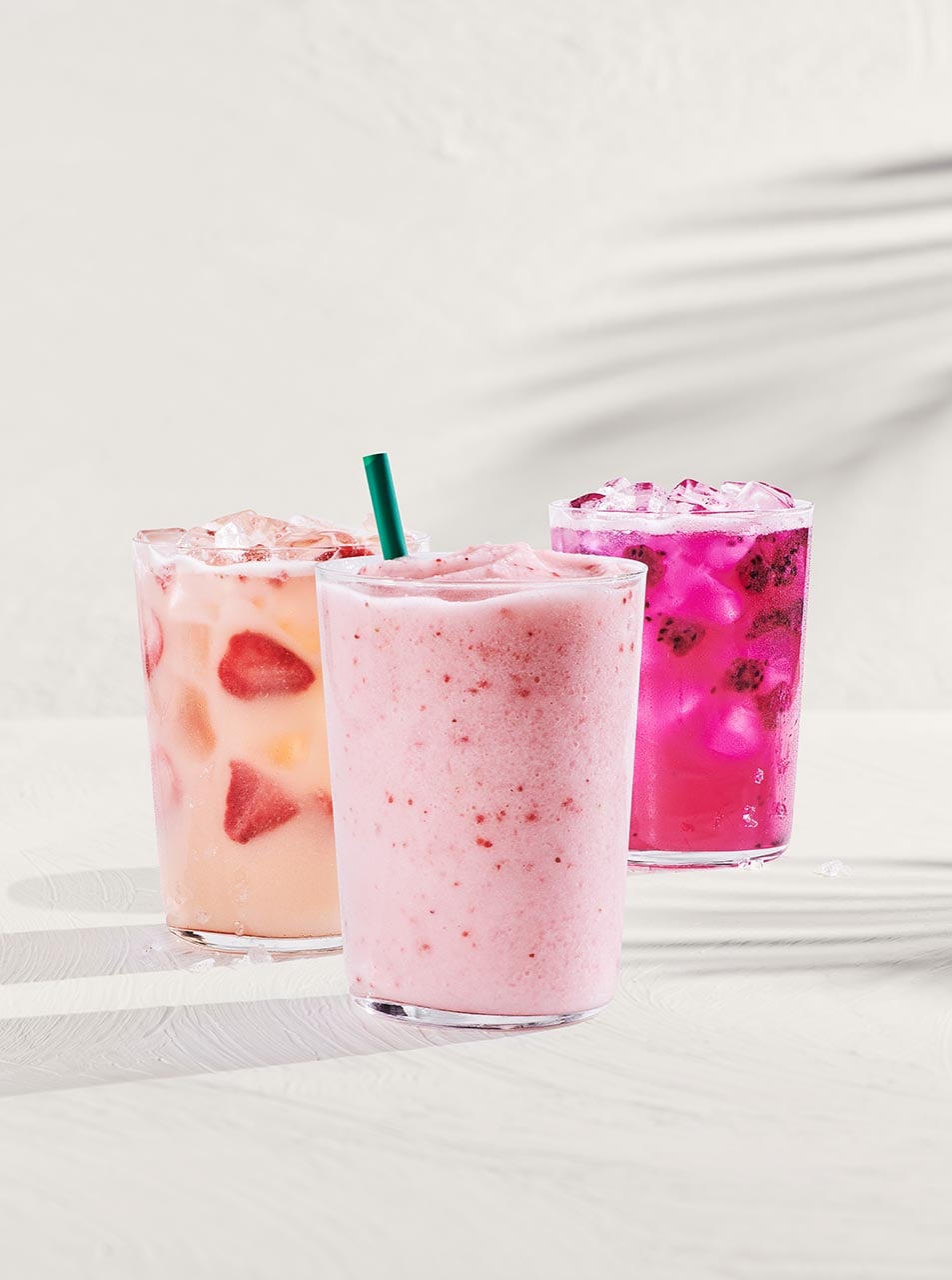 Three colorful drinks are displayed in clear glasses. The left drink is a peach and strawberry mix with ice, the center is a pink smoothie, and the right is a vibrant purple beverage with ice and dark fruit pieces.