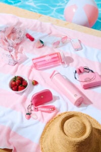 A vibrant pink beach scene featuring a striped towel, a straw hat, strawberries in a bowl, and various pink accessories like a water bottle, phone, and sunglasses.