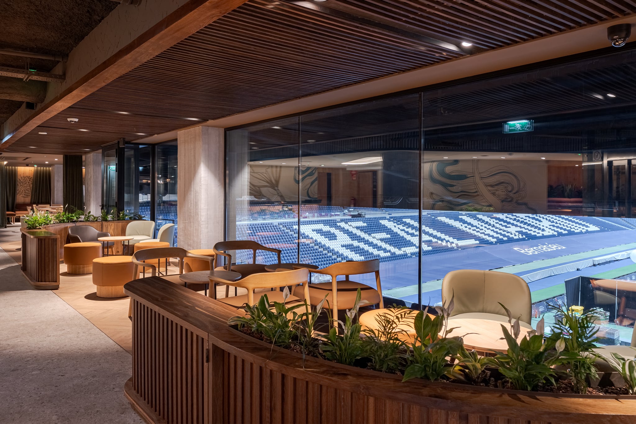 A modern lounge area features wooden furniture and plants, with large glass windows overlooking a sports stadium seating area.
