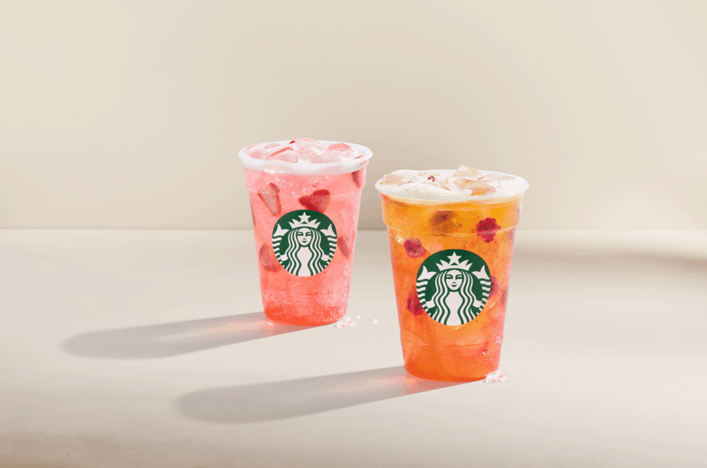 Two clear cups filled with colorful iced beverages are displayed. The left cup contains a pink drink with ice and fruit pieces, while the right cup features a yellow-orange drink topped with cream and fruit. Both cups have the Starbucks logo prominently displayed.