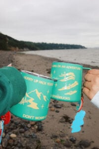 Two colorful mugs are held up against a beach backdrop. The mugs feature adventure-themed graphics and text, with a sandy shore and water in the background.