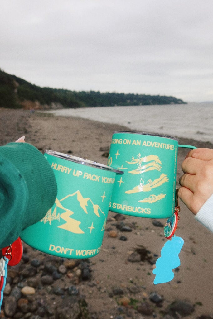 Two colorful mugs are held up against a beach backdrop. The mugs feature adventure-themed graphics and text, with a sandy shore and water in the background.