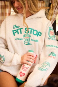 A woman is wearing a light gray hoodie with colorful text and graphics related to a "Pit Stop" theme. She holds a pink and green water bottle featuring "Road Trip!" and "Sunset Drives" text. The background shows a cozy interior with patterned curtains.