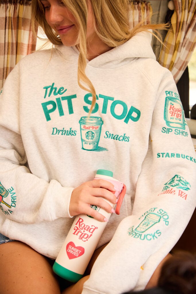 A woman is wearing a light gray hoodie with colorful text and graphics related to a "Pit Stop" theme. She holds a pink and green water bottle featuring "Road Trip!" and "Sunset Drives" text. The background shows a cozy interior with patterned curtains.