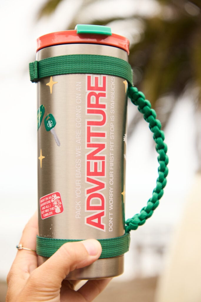 A stainless steel travel tumbler with a red lid and green strap, featuring the word "ADVENTURE" and playful graphics.