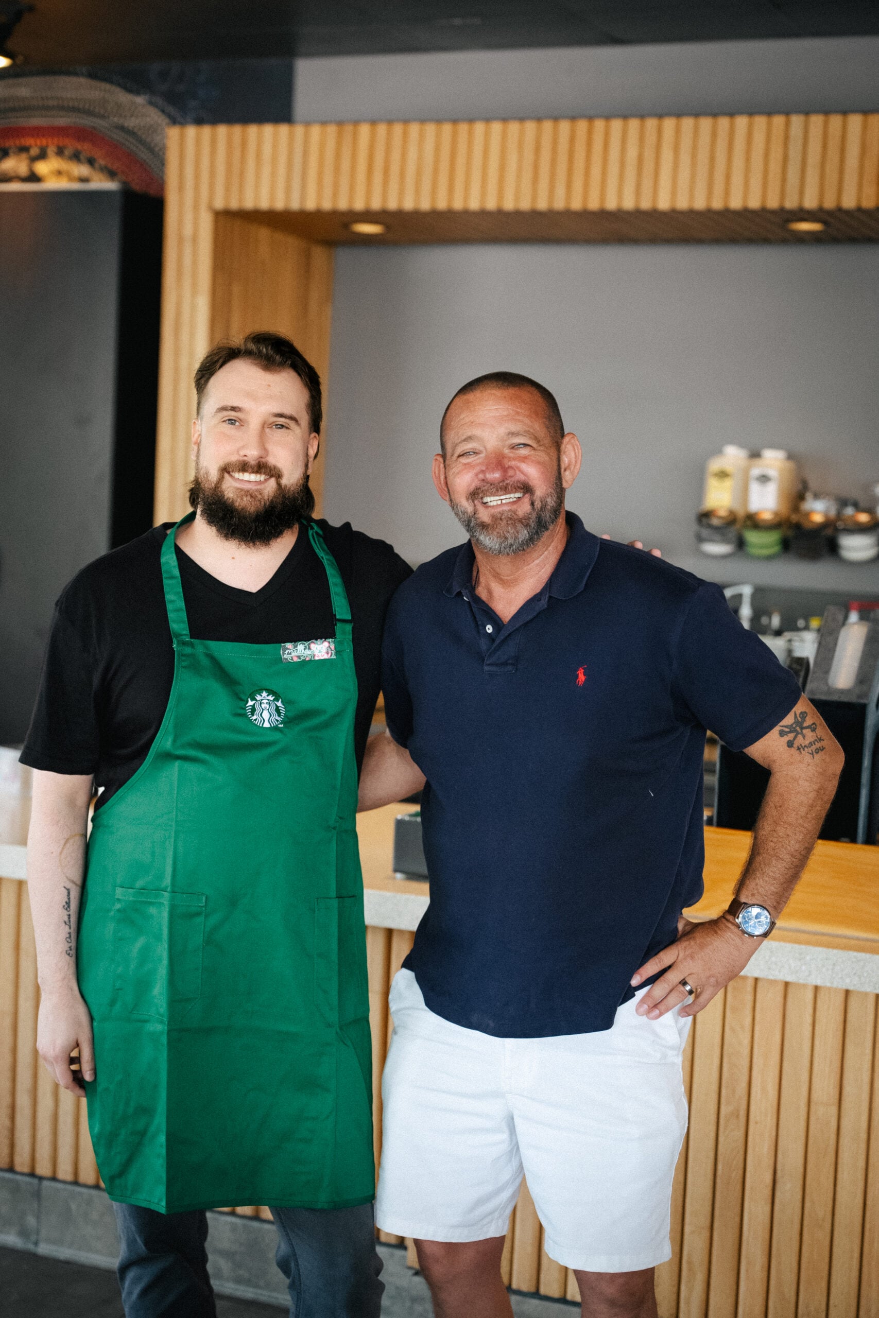 Two men pose together in a café setting. One wears a green apron with a logo, while the other is in a navy polo and white shorts. The background features wooden accents and café equipment.