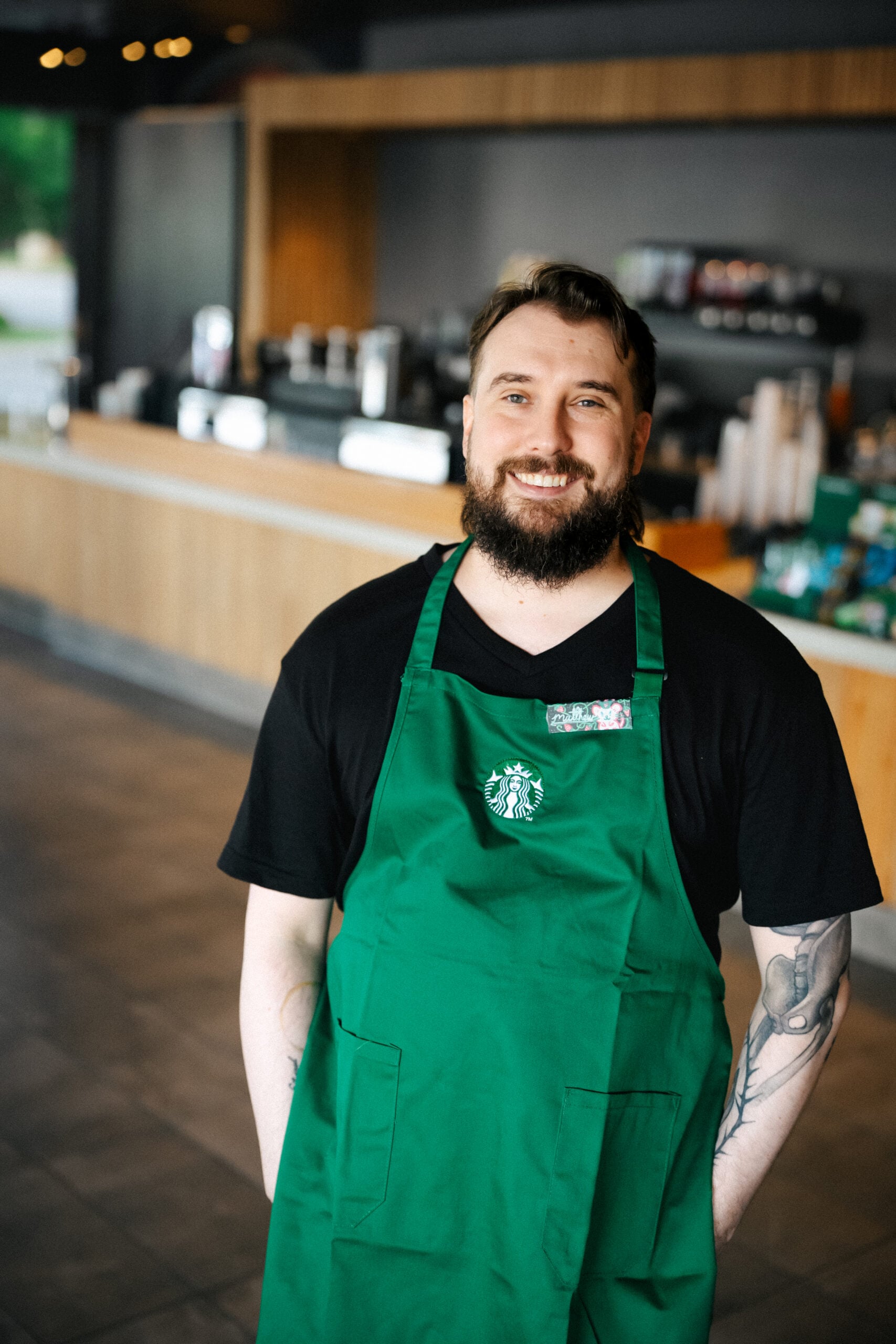 A smiling barista in a green apron stands in a coffee shop, with a modern counter and coffee equipment visible in the background.