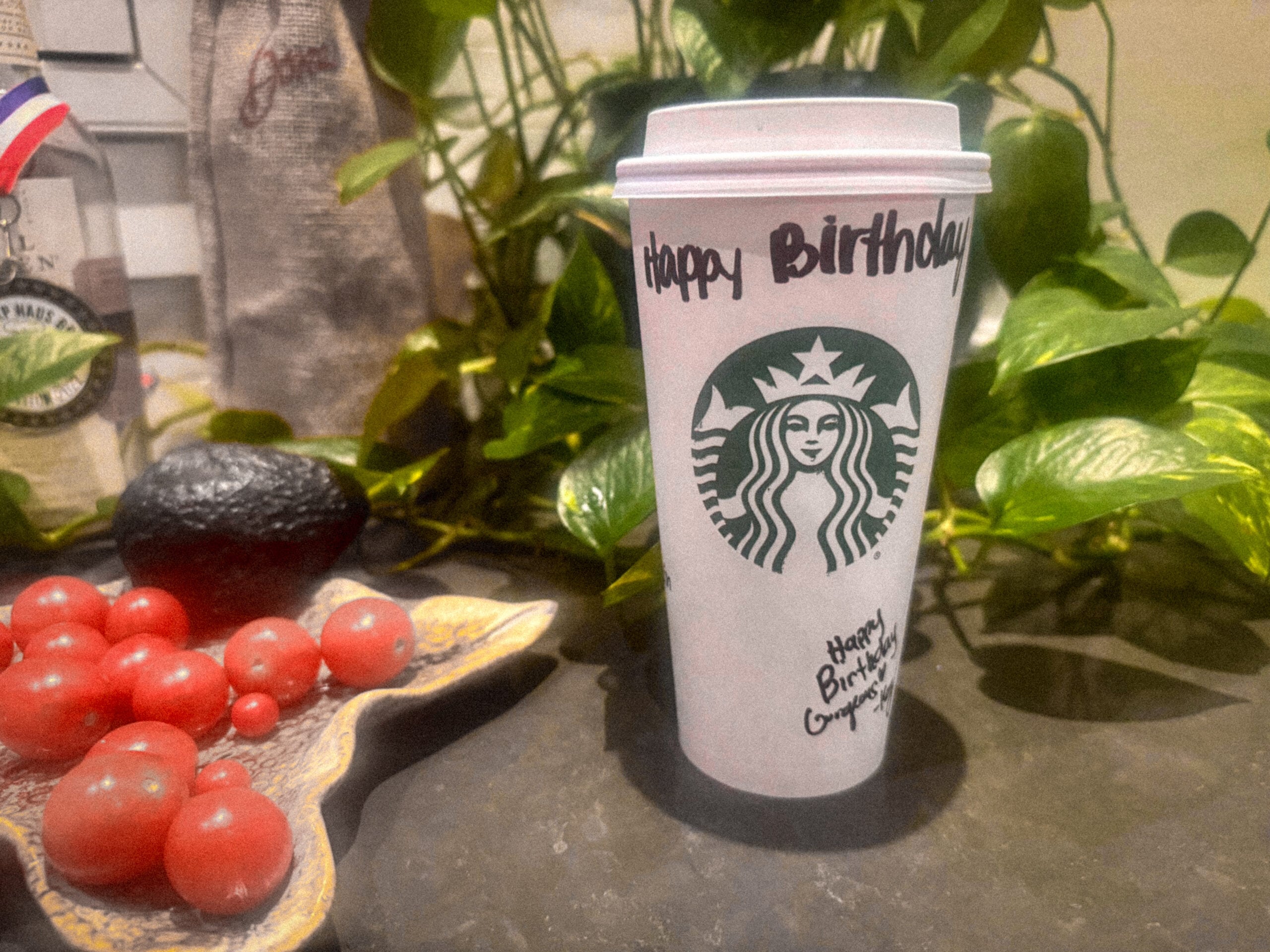 A Starbucks coffee cup with "Happy Birthday" written on it sits on a dark surface. Surrounding it are red cherry tomatoes and an avocado, with green plants in the background.