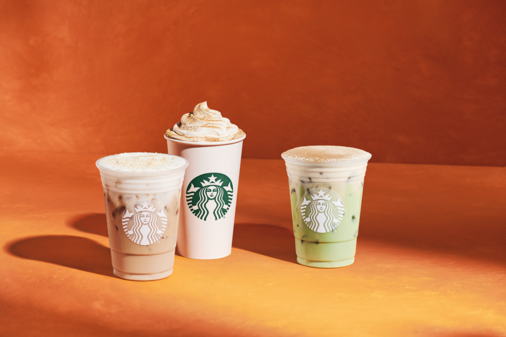 Three Starbucks beverages are displayed against an orange background: an iced drink with a light foam top, a hot drink with whipped cream, and a green iced drink. Each cup features the Starbucks logo.