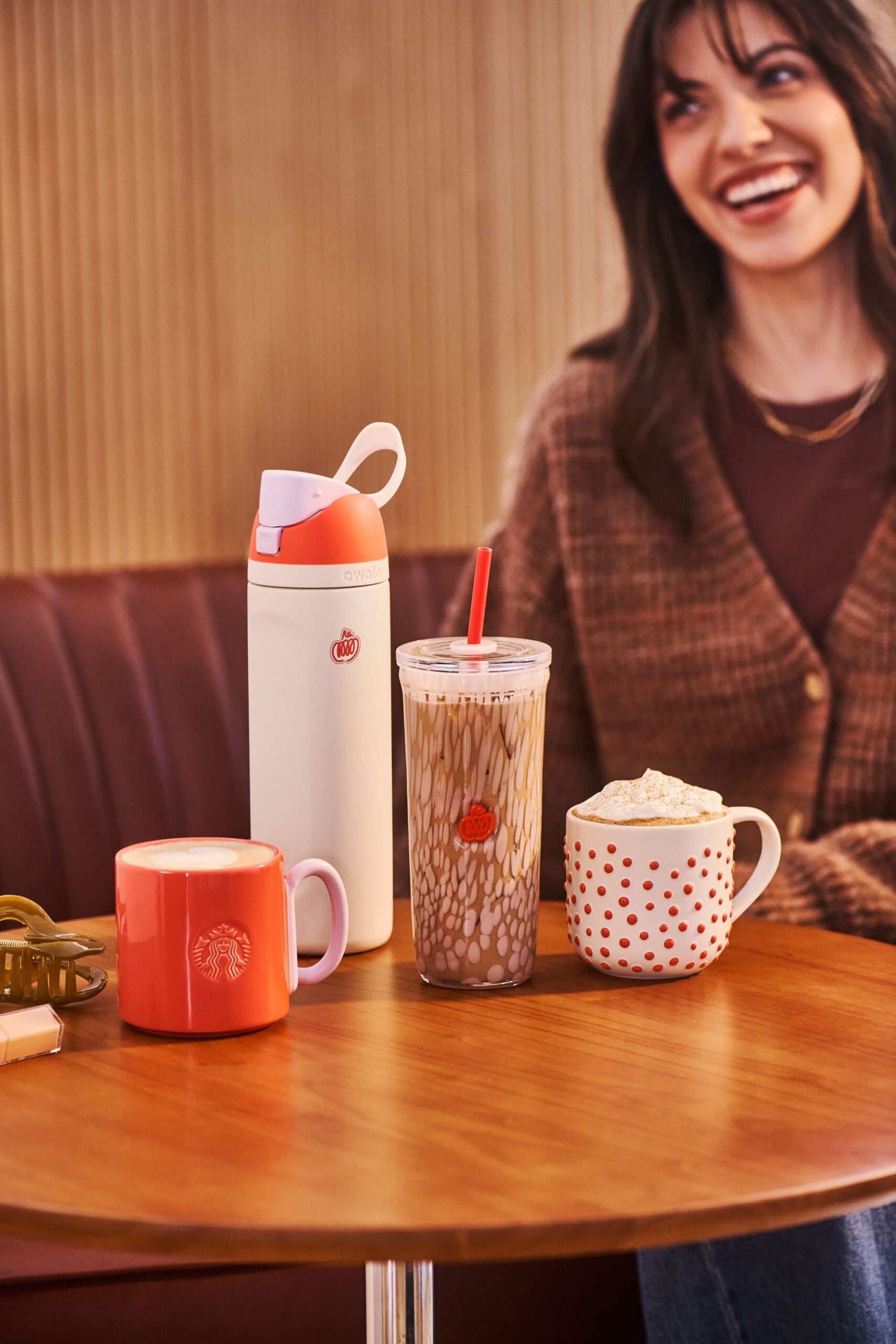 A cozy scene featuring a wooden table with three drink containers: a coral mug, a patterned iced coffee cup, and a white thermos. A smiling woman in a brown sweater is in the background.