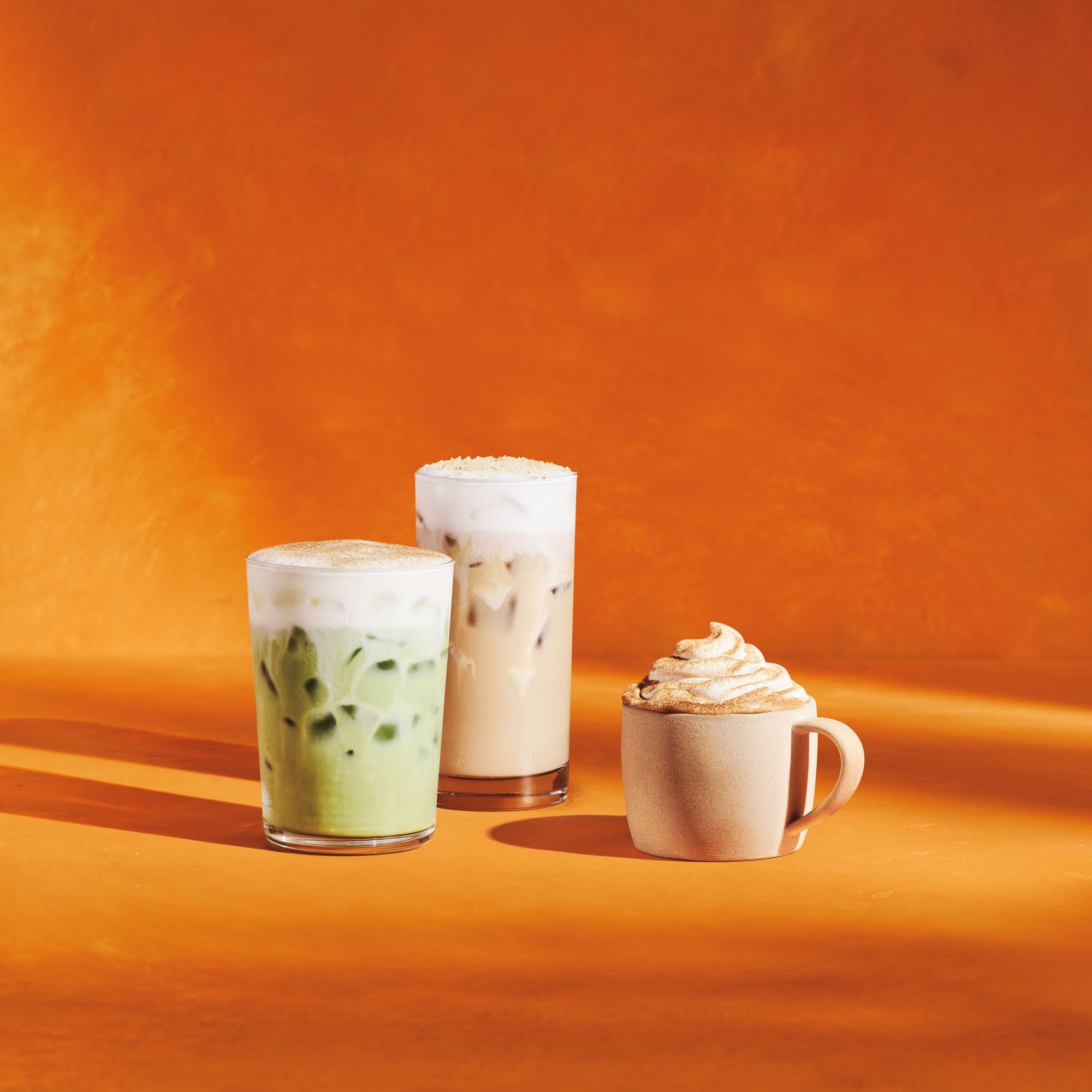Three beverages are displayed against an orange background: a green iced drink with foam, a tall iced coffee topped with foam, and a small cup with whipped cream.