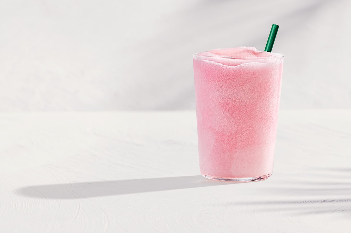 A tall glass filled with a frosty pink beverage topped with a green straw, set against a light background.