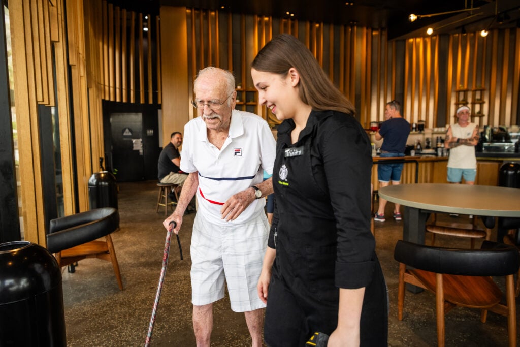 An elderly man with glasses and a cane walks alongside a young woman in a black apron. They are in a modern café with wooden accents.