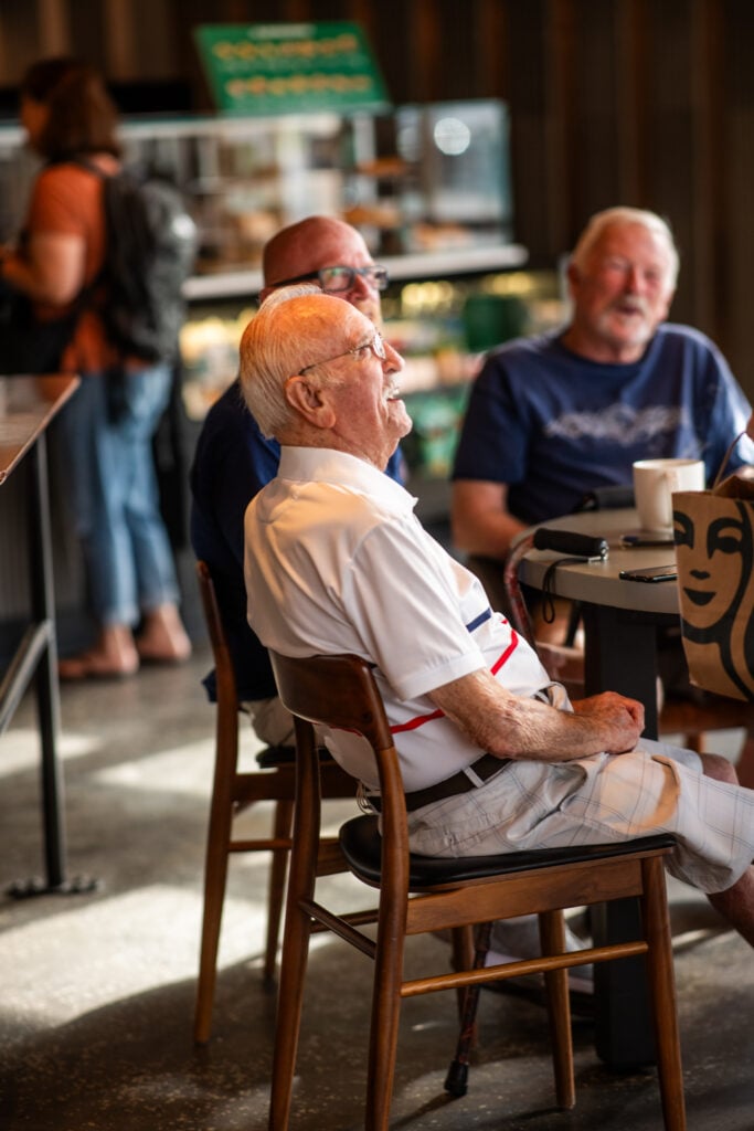 A group of older men sit at a café table, engaged in conversation. One man, wearing a white shirt with red stripes, smiles while seated. The background features a café display and other patrons.