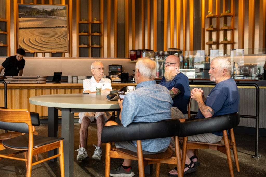 A group of four older men sit around a round table in a cafe. The setting features wooden accents and a modern design. One man in a white shirt appears to be listening intently, while another looks at his phone. The others are engaged in conversation.