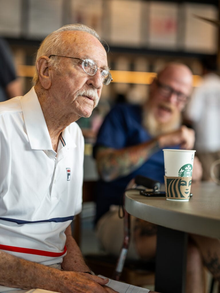An elderly man with glasses sits at a table, wearing a white polo shirt. A Starbucks cup is on the table, with a blurred figure laughing in the background.