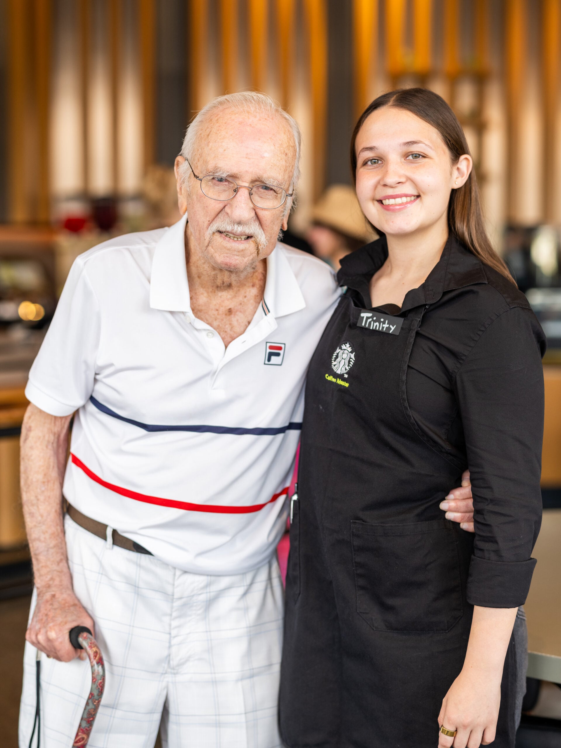 An elderly man in a white polo shirt with red and blue stripes stands next to a young woman in a black apron. They are smiling, with the woman’s name tag visible. The background features a modern café setting.