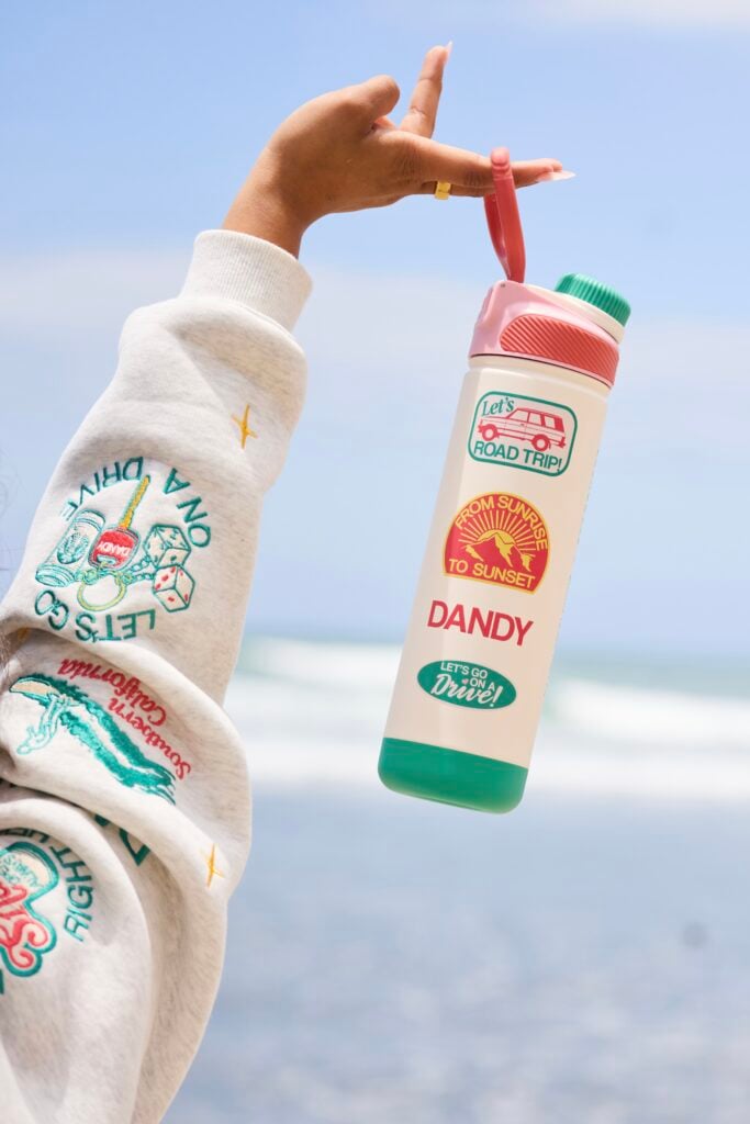 A hand holds a colorful water bottle with travel-themed stickers, against a beach backdrop. The bottle features a pink lid and green base.