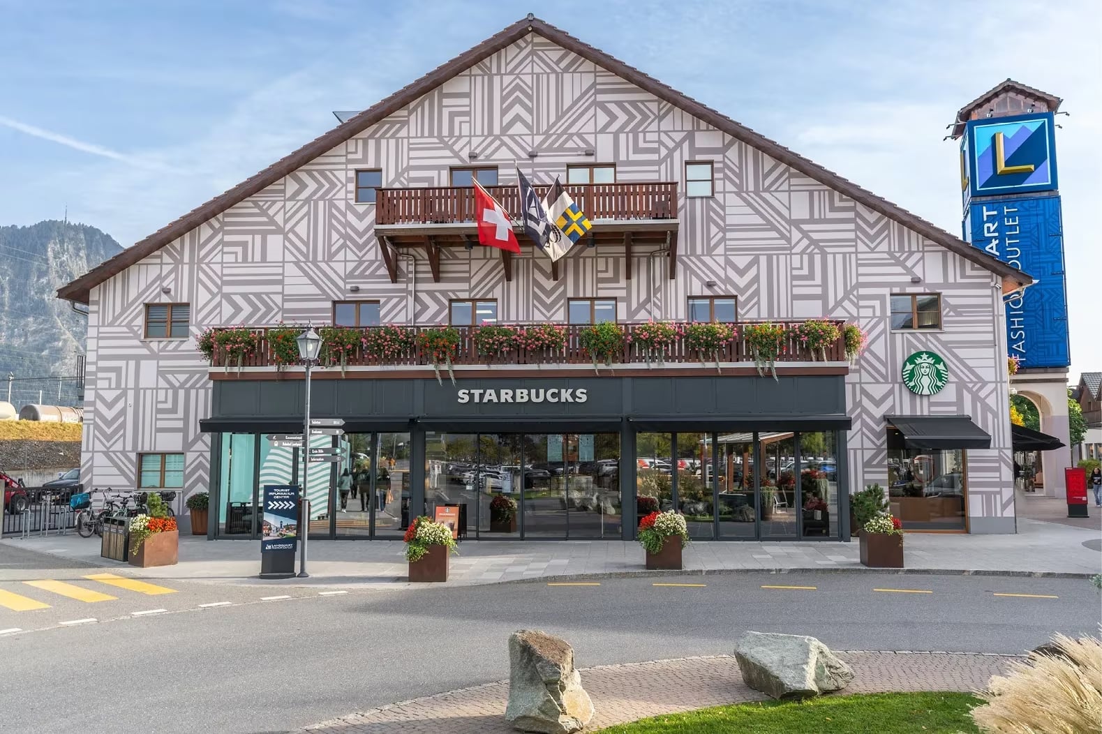 A Starbucks store features a patterned facade with flower boxes and flags. Nearby, a tall sign indicates a fashion outlet.