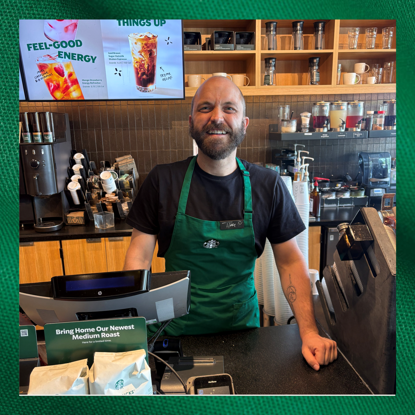 A barista with a beard smiles at the camera while standing behind a coffee shop counter. He wears a green apron over a black shirt. Behind him, a digital menu displays beverages, and various coffee supplies are visible on the shelves.